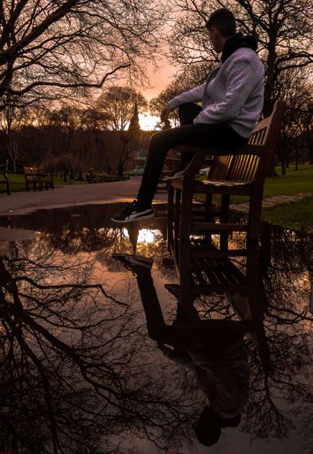A vertical shot of the reflection of a male sitting on a bench on a pond near trees in a park during sunset