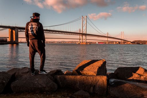 A wide shot of a male wearing a black cap and hoodie standing on a rock by a cable-stayed bridge on the body of water