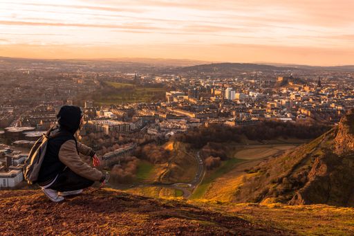 A wide shot of a person sitting at the top of a mountain surrounded by buildings in a city during sunset
