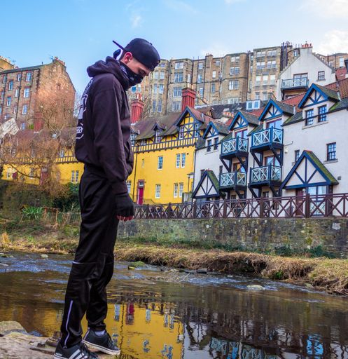 A vertical shot of a person wearing a hoodie and black cap standing at a dirty pond near buildings and houses