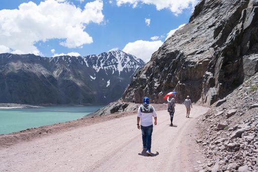 A group of several people walking on a country road next to a turquoise water reservoir with beautiful snowy mountains in the background