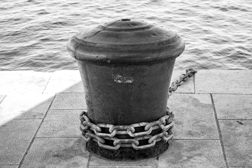 A grayscale shot of a metal mooring bollard at the pier on a sunny day