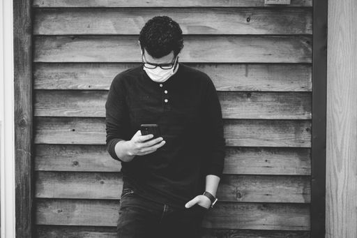 A greyscale shot of a male in glasses and a face mask using his phone in front of a wooden wall