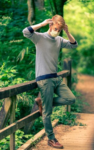 A blonde male wearing a sanitary face mask while standing on a wooden bridge in a forest