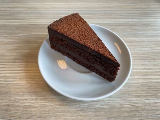 A piece of chocolate cake sprinkled with cocoa on a white plate lying on a wooden table.