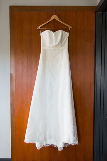 A vertical shot of a long white dress hanging from a cloth hanger on a door under the lights