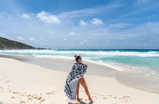 Woman wrapped in beach towel standing on long sandy beach with turquoise ocean and big waves on La Digue island on Seychelles.