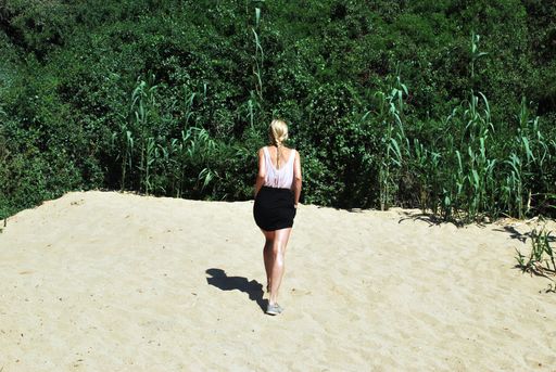 A female walking on a sandy surface towards a grassy field with plants and trees shot from behind at daytime