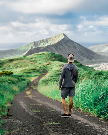 A vertical shot of a young male walking through a pathway surrounded by greenery and mountains under a cloudy sky