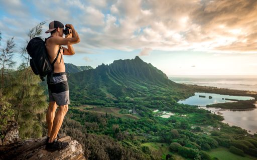 A young shirtless male with a backpack standing on a mountain and taking a picture under a cloudy sky