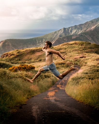 A young attractive shirtless male jumping in a field surrounded by mountains under the sunlight and a cloudy sky