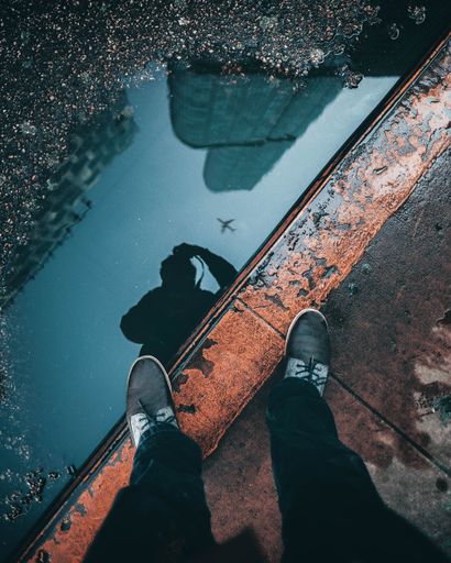 A vertical overhead shot of the reflection of a person and buildings in a water puddle