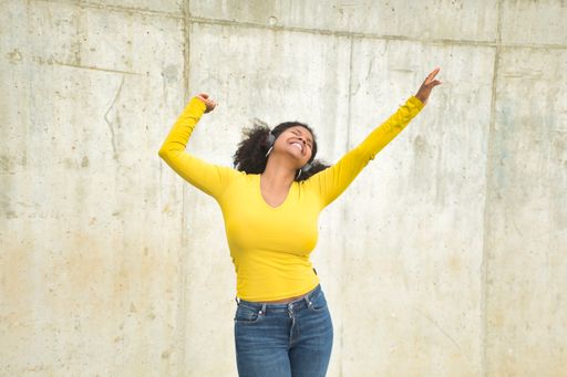 A closeup of an African American woman with afro hair, a yellow t-shirt, and Bluetooth headphones dancing happily