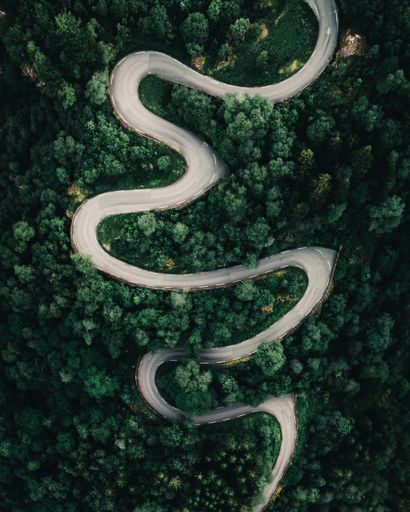 A vertical drone shot of a winding road passing through forests with a rapidly flowing waterfall next to the road