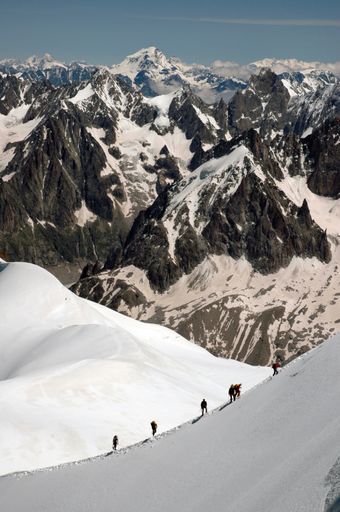 A vertical shot of hikers hiking a snowy hill surrounded by mountains covered with snow