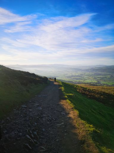 A hillside path with a man walking toward the foggy scenery in the distance on a sunny day in Wrexham