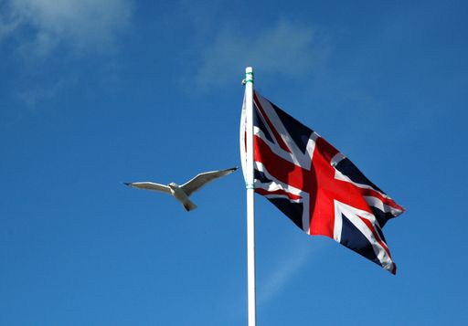 A white bird flying in a blue sky near a flagpole with the United Kingdom flag