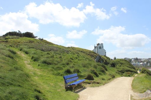 A  beautiful shot of a blue chair near the grassy hill with a building in the distance under a cloudy sky