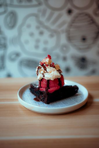 A Close-up shot of a rich chocolate brownie with melted strawberry ice cream, chocolate sauce, cream and red candy sprinkles in a white plate