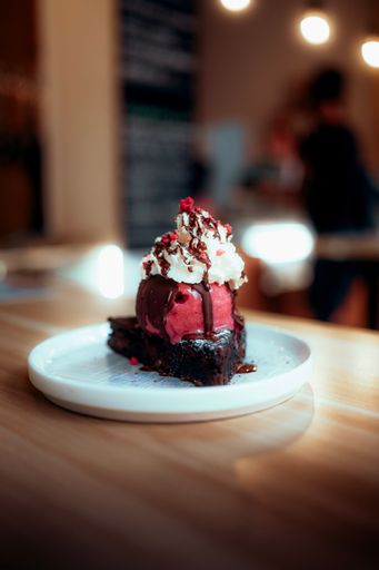 A Close-up shot of a rich chocolate brownie with melted strawberry ice cream, chocolate sauce, cream and red candy sprinkles in a white plate