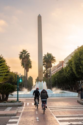 The Two tourists on bikes at the sculpture and water fountain on the Belen street of the Rambla de Almeria at sunset, Andalucia. Spain.