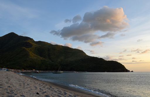 A beautiful shot of a beach shore with a boat in the distance near a forested mountain under a blue sky