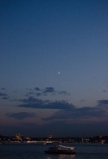 A vertical shot of a boat on the body of water in a city under a night sky with a half moon