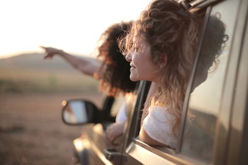 A curly-haired woman pointing her hand out of the car window next to a blonde lady