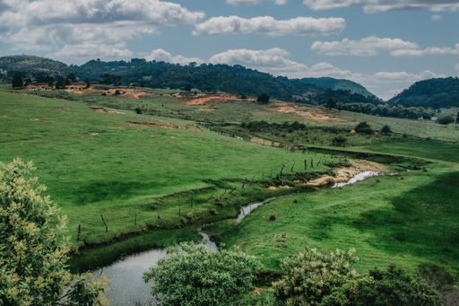 A beautiful view of a river flowing through the field with green grass with the background of the mountains under the cloudy sky