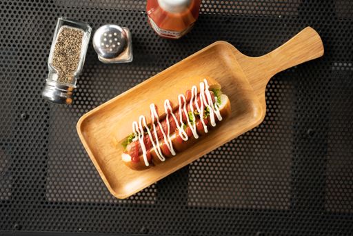 An overhead shot of a hotdog with sauce on a wooden tray near salt and pepper
