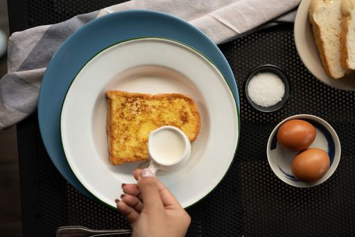 An overhead shot of a person pouring cream on top of a french toast near eggs and sugar