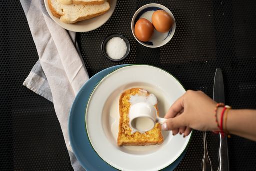 An overhead shot of a person pouring cream on a french toast near eggs and sugar
