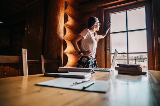 A young female in a white shirt standing next to a window in the wooden cabin with a working table