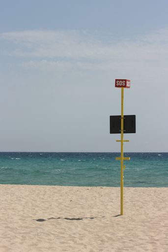 A vertical shot of an SOS sing on a sandy surface near the water with a clear sky in the background