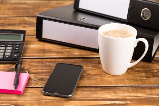 A cup of coffee, smartphone, folder, calculator, pen, and a sticky note on a desk