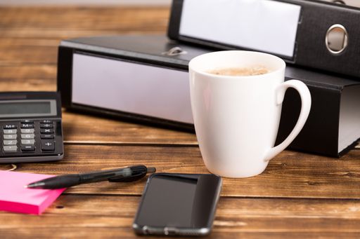 A cup of coffee, smartphone, folder, calculator, pen, and a sticky note on a desk