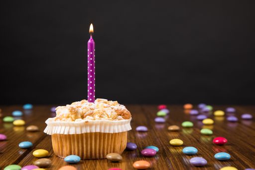A cupcake with a burning candle, and colorful candies on the wooden table against a black wall