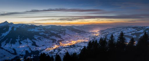 A panoramic shot of buildings with lit lights near snowy mountains under a blue sky at night time