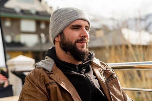 A profile shot of a bearded man in a hat in the background of some buildings.