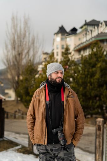 A vertical shot of a young bearded guy standing in the background of buildings and spruces in winter.