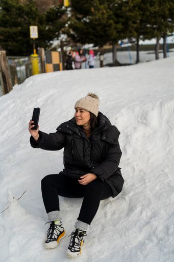 A vertical shot of a young Caucasian woman sitting on the snow and taking a selfie.