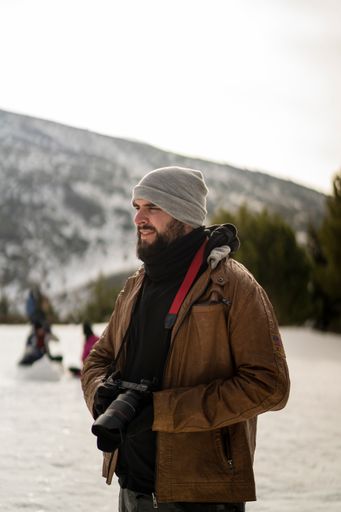 A vertical shot of a young bearded guy with a camera standing in the background of nature.