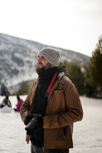 A vertical shot of a young bearded guy standing in the background of mountains and spruces in winter.