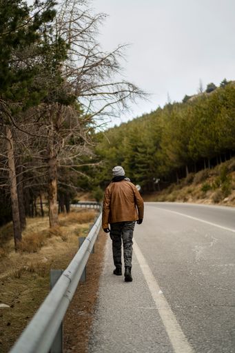 A vertical shot of a young guy walking on the edge of the highway in the background of trees.