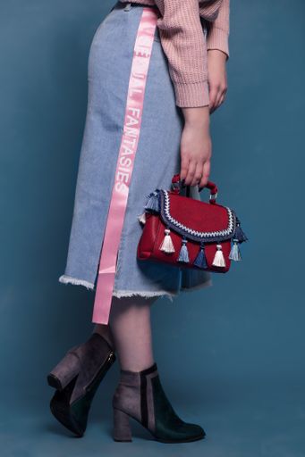 A vertical shot of a woman in a blue long skirt standing near a blue wall and holding a red bag