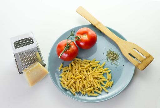 A high angle shot of Italian organic pasta with tomatoes, seasonings, and a wooden spoon in a blue plate next to cheese and grater