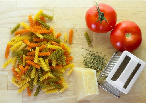 A high angle shot of a chopping board with a grater, cheese, Italian colored pasta and tomatoes on a white background