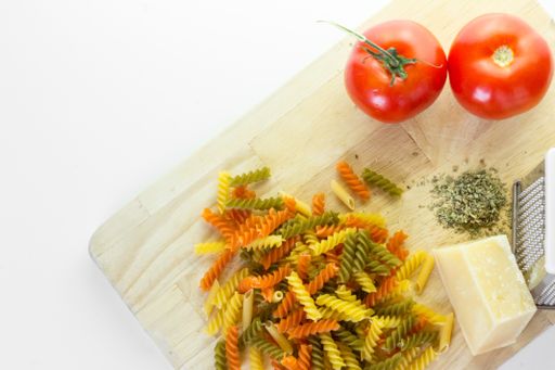A high angle shot of a chopping board with a grater, cheese, Italian colored pasta and tomatoes on a white background