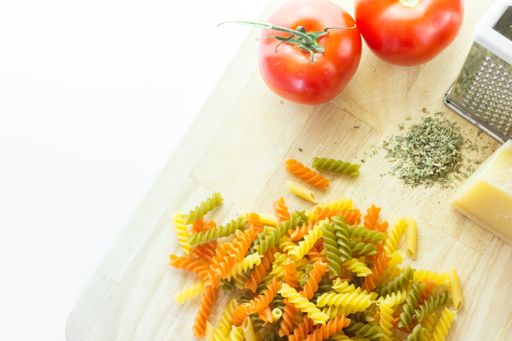 A high angle shot of a chopping board with a grater, cheese, Italian colored pasta and tomatoes on a white background