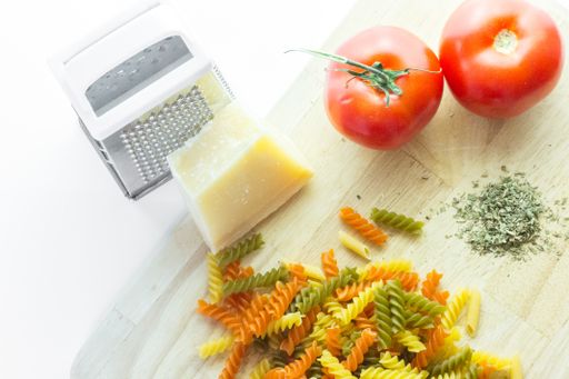 A high angle shot of a chopping board with a grater, cheese, Italian colored pasta and tomatoes on a white background
